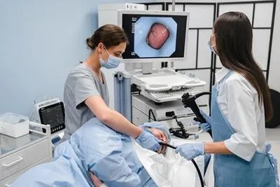 Equipe médica composta por duas profissionais de saúde realizando um exame de colonoscopia em um paciente. Uma médica manipula o endoscópio enquanto a outra auxilia no posicionamento, com um monitor ao fundo exibindo a imagem interna do cólon.