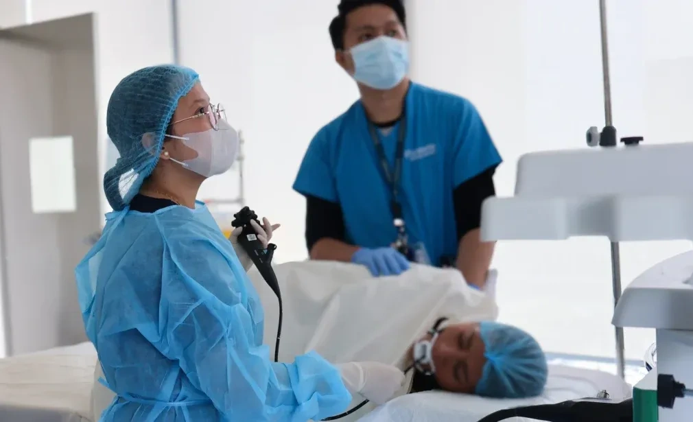 Profissionais de saúde em trajes cirúrgicos azuis, máscaras e toucas, realizando um exame de colonoscopia. Uma médica opera o equipamento enquanto a equipe monitora atentamente o procedimento em um ambiente clínico iluminado.